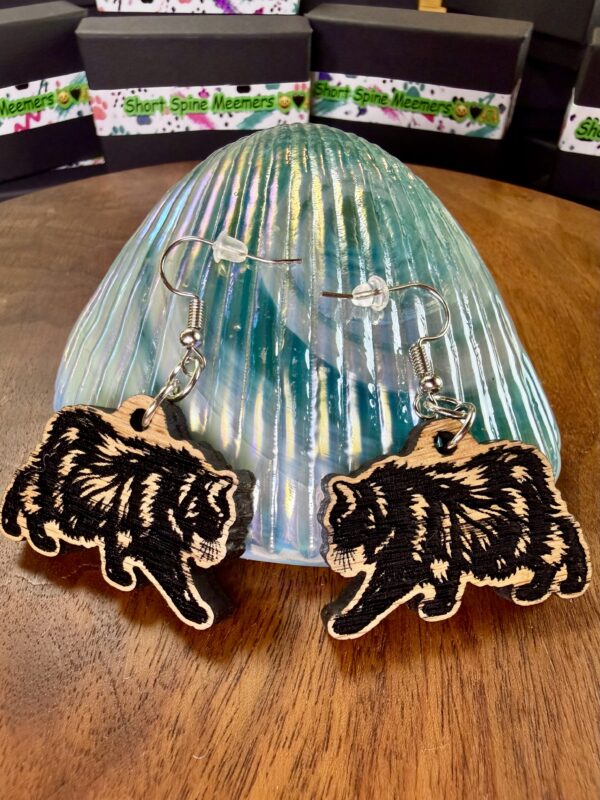 Pair of red oak Meemers dangle earrings with black engraved cat designs on an iridescent teal stand